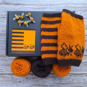 May include: An orange and brown knitted leg warmer set with a bee design, next to a box with bee-themed decorations and three balls of yarn. The leg warmers have a striped pattern and bee graphics.