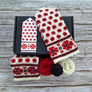 May include: A set of white and red knit mittens with a poppy flower pattern. The mittens are packaged in a black box with a white label that shows the pattern. The set also includes three balls of yarn in red, black, and white.