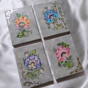 May include: Four rectangular canvases with a textured gray background, each featuring a hand-painted floral design. The flowers are rendered in vibrant shades of orange, blue, purple, and pink, accented with green leaves and metallic gold or silver swirls.