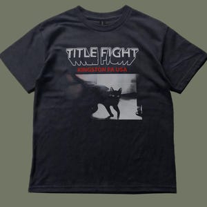 May include: A black short-sleeve t-shirt featuring the white text "TITLE FIGHT" above the red text "KINGSTON PA USA". Below the text is a rectangular graphic of a black cat standing in a room with a light-colored floor and dark shadows.