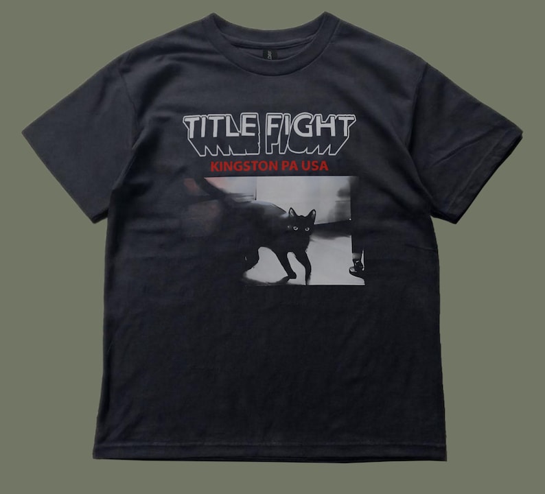 May include: A black short-sleeve t-shirt featuring the white text "TITLE FIGHT" above the red text "KINGSTON PA USA". Below the text is a rectangular graphic of a black cat standing in a room with a light-colored floor and dark shadows.