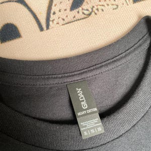 May include: A black crew neck t-shirt featuring a ribbed collar and a black fabric tag attached to the inner neckline. The tag displays the brand name GILDAN, the text HEAVY COTTON, and the size XL, TG, EG printed in white lettering.