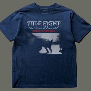 May include: A navy blue short-sleeve t-shirt featuring the white text "TITLE FIGHT" above the red text "KINGSTON PA USA". Below the text is a black and white photographic print of a cat standing in a room with visible furniture legs.