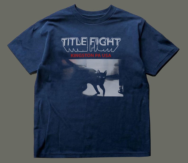 May include: A navy blue short-sleeve t-shirt featuring the white text "TITLE FIGHT" above the red text "KINGSTON PA USA". Below the text is a black and white photographic print of a cat standing in a room with visible furniture legs.