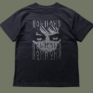 May include: Dark grey t-shirt with a graphic print of a face and the word "BAUHAUS" repeated vertically. The design is in shades of grey. The t-shirt has a crew neck and short sleeves.
