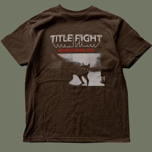May include: A brown short-sleeve t-shirt featuring the text "TITLE FIGHT" in white and "KINGSTON PA USA" in red. Below the text is a black and white graphic of a cat walking across a floor, with the cat's eyes clearly visible in the image.