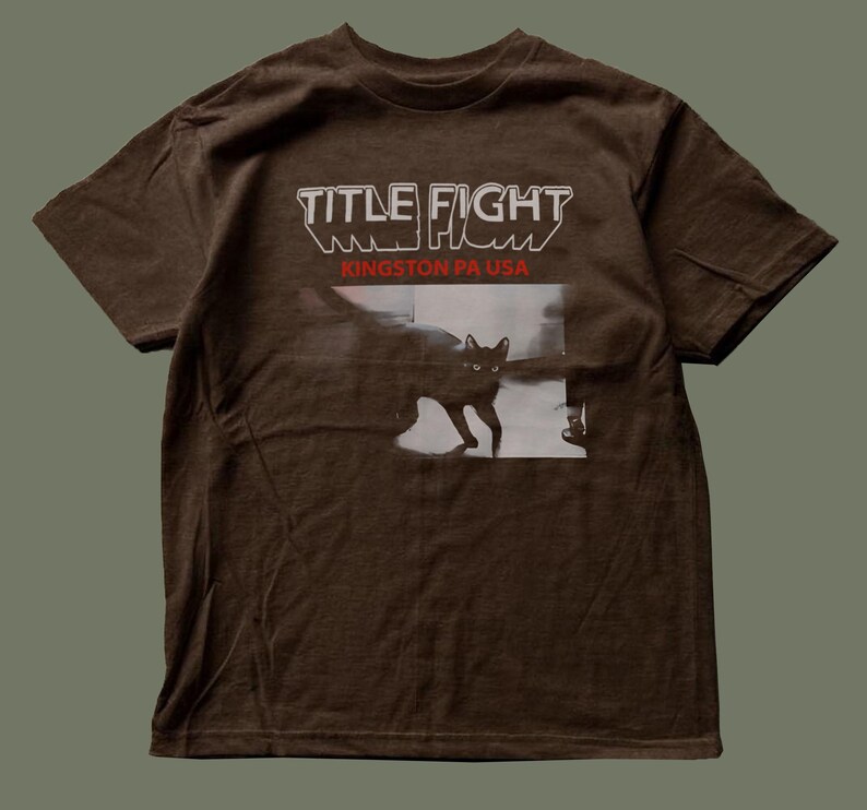 May include: A brown short-sleeve t-shirt featuring the text "TITLE FIGHT" in white and "KINGSTON PA USA" in red. Below the text is a black and white graphic of a cat walking across a floor, with the cat's eyes clearly visible in the image.