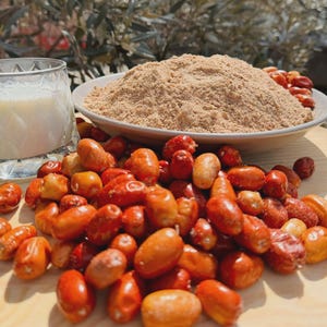 May include: A white bowl filled with a mound of light brown powder, surrounded by a large pile of small, oval-shaped orange and red fruits. A clear glass filled with white liquid sits to the left of the bowl on a light wooden surface.