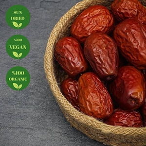 May include: A woven basket filled with sun-dried red dates. The basket is on a gray background. The labels on the left side of the image read "Sun Dried", "100% Vegan", and "100% Organic".