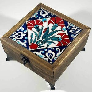 May include: A square wooden box with a decorative tile top. The tile features a floral design with red flowers, teal leaves, and blue and white accents. The box has a dark wood finish and black metal feet.
