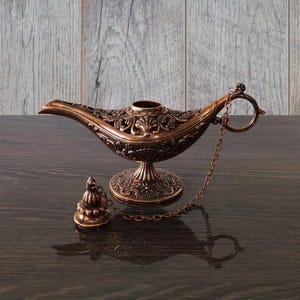 May include: Ornate bronze-colored genie lamp with intricate floral and scrollwork detailing. The lamp features a long spout, a round base, and a small lid attached by a chain. The lamp is a decorative item.