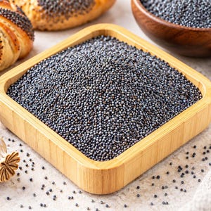 May include: A square wooden bowl filled with small, dark blue-gray poppy seeds. The bowl is positioned near a baked roll topped with poppy seeds, a wooden bowl of seeds, a small wooden spoon, and two dried poppy seed pods.