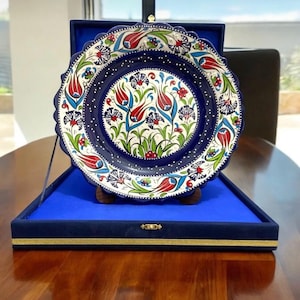May include: A decorative ceramic plate with a scalloped edge, featuring a vibrant floral design in red, blue, and green against a white background. The plate is displayed in a blue velvet presentation box with a gold trim.
