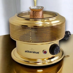 May include: A gold-colored electric coffee warmer with a copper coffee pot and a gold spoon. The warmer has a perforated design and a control knob. The brand name "albarakat" is printed on the front.