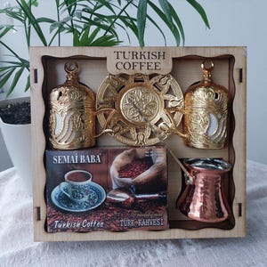 Può includere: Un set da caffè turco in una scatola di legno. Il set include due tazze da caffè dorate ornate, un piatto da portata dorato, una caffettiera in rame e una scatola di caffè turco. La scatola è etichettata "Turkish Coffee".
