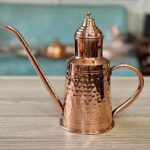 May include: A copper watering can with a long, slender spout and a hammered texture. The can has a handle and a decorative lid with a small knob. The copper finish gives it a warm, rustic appearance. The watering can is designed for indoor or outdoor use.