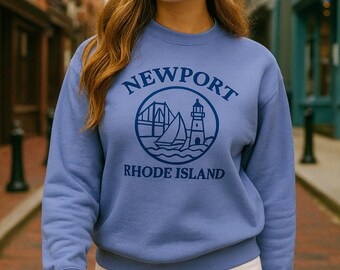 May include: A periwinkle blue sweatshirt with the word "NEWPORT" arched above a circular graphic of a bridge, sailboat, and lighthouse. Below the graphic is the text "RHODE ISLAND". The sweatshirt is crew neck and long-sleeved.