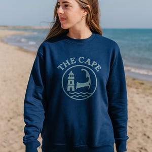 Op de afbeelding: Marineblauwe sweatshirt met een cirkelvormige afbeelding met een vuurtoren, golven en de tekst "THE CAPE". Het ontwerp is in een lichtere blauwtint. De sweatshirt wordt op het strand gedragen.