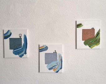 May include: Three square abstract paintings on a white wall. Each canvas features a white background with geometric shapes and brushstrokes in blue, green, and brown. One painting includes a hot air balloon, another a sailboat.