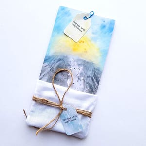 May include: A watercolour painting of a sunset over a body of water. The painting is wrapped in a white cloth with a tag that reads "Journey with confidence".