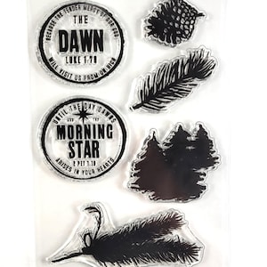 May include: Clear sheet with six black and white rubber stamps. The stamps feature a pine cone, a pine branch, a pine tree, a feather, and two circular stamps with religious text. The text on the stamps reads "The Dawn Luke 1:78 Will Visit Us From On High" and "Until The Day Dawns And The Morning Star Arises In Your Hearts 2 Pet 1:10".