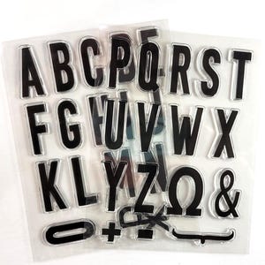 May include: Clear plastic sheet with black alphabet letter stamps. The letters include A, B, C, D, E, F, G, H, I, J, K, L, M, N, O, P, Q, R, S, T, U, V, W, X, Y, Z, &, and 0.