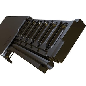 Op de afbeelding: Een zwart metalen gereedschap met meerdere gebogen sleuven en een cilindrisch onderdeel. Het gereedschap heeft een rechthoekige vorm met een schuine bovenkant. Het ontwerp is industrieel en functioneel.