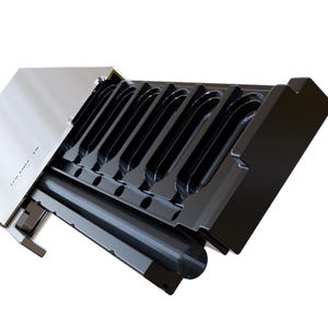 May include: A black and silver metal tool with a series of curved slots. The tool has a rectangular shape with a hinged lid. The slots are designed to hold a specific item. The tool is angled, revealing its interior.