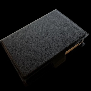 Puede incluir: Un cuaderno negro rectangular encuadernado en cuero con una superficie texturizada. El cuaderno está cerrado, con una pestaña visible. El fondo es negro liso.