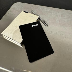 Puede incluir: Una tarjeta rectangular negra con el texto "el patron" en letras blancas descansa sobre una superficie metálica. Al lado, una caja rectangular beige con el mismo texto. También hay un bolígrafo plateado.