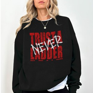 Op de afbeelding: Zwarte sweatshirt met de tekst "TRUST A NEVER LADDER" in rode en witte letters. De zin "They're always up to something" staat onder de hoofdtekst.