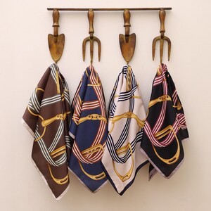May include: Four silk scarves with a gold chain pattern on a white, navy blue, brown, and black background hanging on a rustic metal rack.