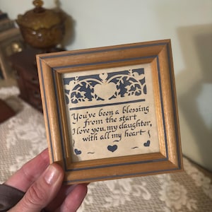 May include: A small, square wooden picture frame with a blue border. The framed artwork features a cutout design and the text: "You've been a blessing from the start, I love you, my daughter, with all my heart."