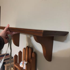 May include: A dark brown wooden shelf with two decorative brackets. The shelf is being measured with a tape measure. The shelf is approximately 30 inches long.
