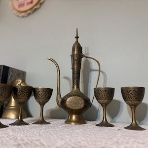 May include: An antique brass tea set with an ornate design. The set includes a tall, slender pitcher with a curved spout and handle, and five small goblets. The pitcher and goblets have a dark, etched pattern.