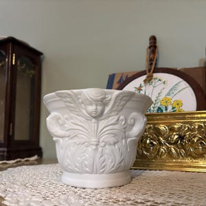 May include: A white ceramic planter with an angel design. The planter features a detailed relief of an angel with wings and decorative scrollwork. The planter is likely for small plants or flowers and is a decorative item.