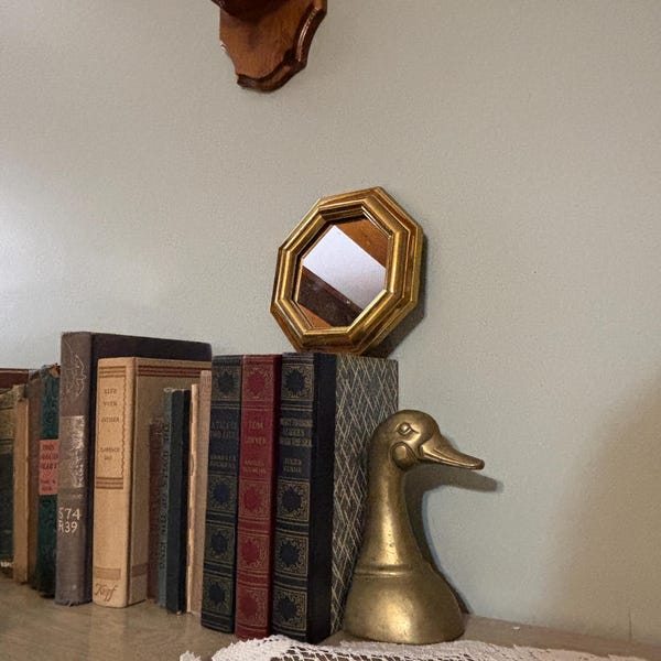 Puede incluir: Una colección de libros antiguos, una figura de pato de latón y un espejo octogonal con marco dorado se exhiben en una estantería. Los libros son de varios colores y tamaños. Una lámpara de pared de madera es visible en el fondo.