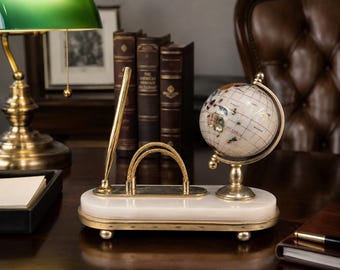 May include: A desk set featuring a white marble base with a gold-toned trim and four small feet. The set includes a gold-toned pen in a vertical holder, a gold-toned arched card holder, and a small rotating globe with a gold-toned metal stand.
