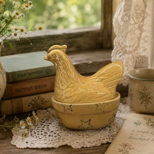 May include: A yellow ceramic chicken-shaped lid sits atop a matching yellow bowl decorated with dark star outlines. The bowl and lid set features a glossy finish and is positioned among various books and floral-themed ceramic items.
