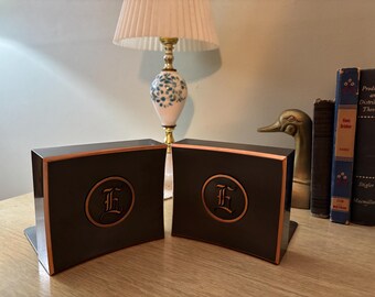 May include: Two black bookends with copper-colored trim and a circular emblem featuring the letter "E". The bookends are on a wooden surface, with a lamp and books in the background. The bookends are designed to hold books upright.