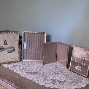 May include: A collection of four metallic photo frames in various sizes. One frame is 8x10 inches, one is a vertical rectangular frame, one is a double-hinged folding frame, and one is a 5x7 inch frame. Each frame features a thin, textured border.