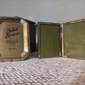 Puede incluir: Dos marcos de fotos de latón. Uno es un marco octogonal con el texto "Solid Brass PHOTO FRAME" y dimensiones de 12,7 cm x 17,8 cm. El otro es un marco con bisagras de doble panel con respaldo verde.