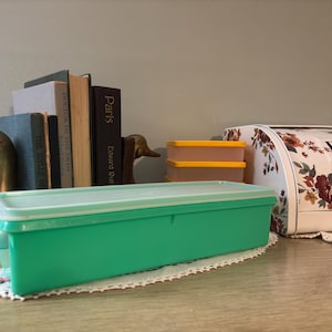 Puede incluir: Un recipiente rectangular verde con tapa blanca sobre un tapete blanco. Dos recipientes naranjas más pequeños apilados detrás. Una panera floral y una colección de libros también son visibles.