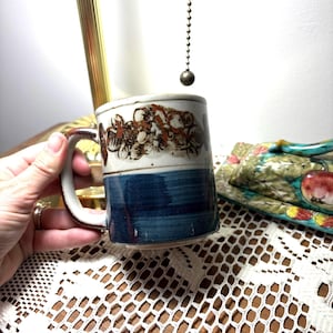 May include: A ceramic mug with a brown and white floral design on the top half and a blue band on the bottom. The mug has a brown handle and is held by a hand. The mug is on a lace tablecloth.