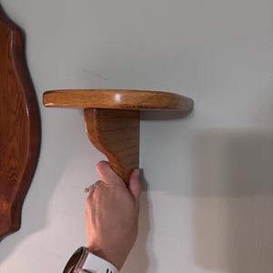 May include: A small, semi-circular wooden wall shelf featuring a smooth, light brown grain finish. The shelf is supported by a single, curved wooden bracket attached to the underside, providing a stable platform for display purposes.