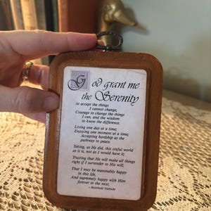 May include: Wooden framed plaque with the Serenity Prayer printed on a white background. The frame is a warm brown color, and the text is in a classic serif font. A decorative metal loop is at the top for hanging.