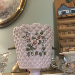 May include: A pale pink, footed vase with a diamond pattern and scalloped rim. The vase is adorned with raised floral details, including pink roses, blue forget-me-nots, and green leaves. A gold band accents the base.