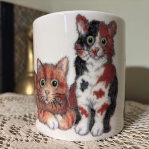 May include: A white ceramic mug featuring two illustrated cats. One cat is orange and the other is calico with black, red, and white markings. The mug is cylindrical and sits on a lace tablecloth.