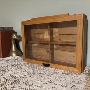 May include: A rectangular wooden display cabinet with a glass front and four interior compartments. The cabinet is made of light-colored wood and has a black handle. A brass duck figurine and books are visible in the background.