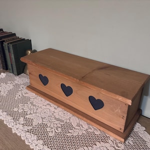 May include: A wooden box with three heart-shaped cutouts. The box is made of light brown wood and sits on a decorative lace doily. A row of books is visible in the background.